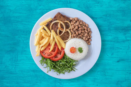 Brazilian food dish. Beans, rice, meat, chips, cabbage, tomatoes, onions. Executive dish. Dish done. Top view. Copy space. Isolated on blue backgroundの写真素材