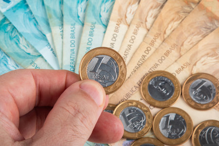 Hand holding Brazilian one Real coin over spread banknotes and coins of Brazilian currency.の写真素材