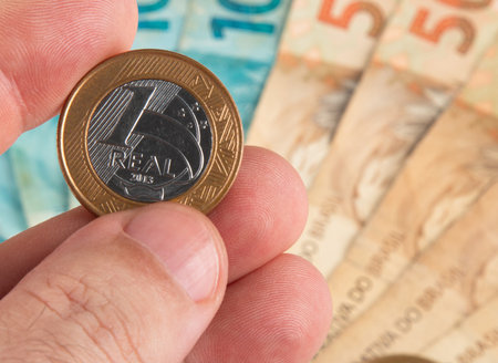 Brazilian one real coin held between fingers with banknotes in the background.の写真素材