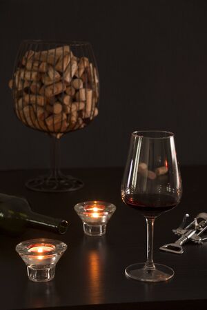 Glass of wine in the foreground with an empty bottle, a corkscrew and corks from the bottles inside a vase simulating a glass of wine in dark tones backgroundの写真素材