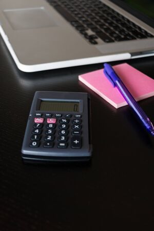 Partial view of a silver keyboard of a laptop with a calculator, and a pink notes with a pencil on a dark wood deskの写真素材