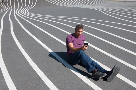 Man reading his smartphone sitting on a grey floor with white stripesの写真素材