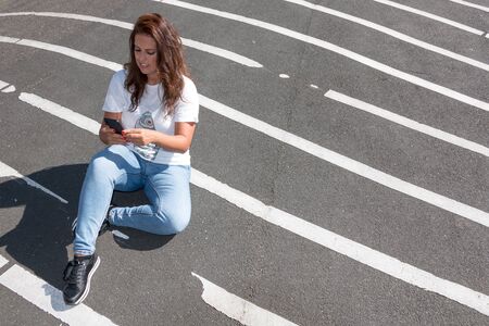 Woman texting on her phone sitting on the asphalt with white lines.の写真素材