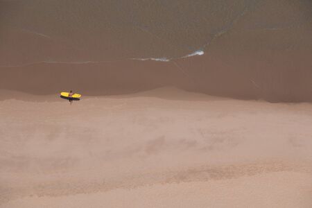 Aerial view of a sandy beach (Praia Cordoama) in Portugal with a surfer and his yellow board heading to the water. Surf conceptの写真素材