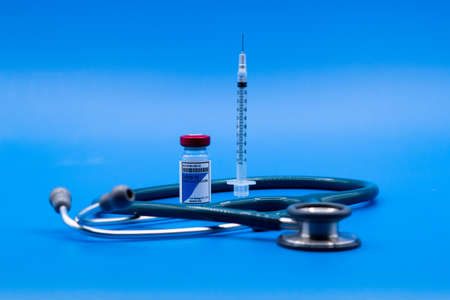 A vial of COVID-19 vaccine with an upright syringe surrounded by a stethoscope on a blue background Medical conceptの写真素材