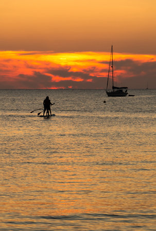 Rear view of two people silhouette on the same board of paddle surf at sunset with a sailboat in background. Holiday conceptの写真素材