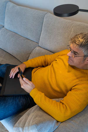 Man sitting on sofa at home texting on phone with closed laptop. Resting at homeの写真素材
