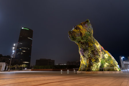 Bilbao, Spain - April 15, 2022: Entrance square to the Guggenheim museum in Bilbao with Puppy, the sculpture of a giant dog made of flowers by Jeff Koons with the Iberdrola tower in the background at nightのeditorial素材