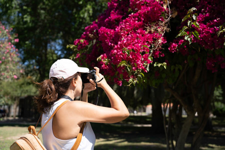 Young woman with a cap and backpack takes a photo with a reflex camera of purple flowers on a tree...