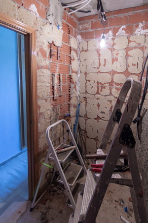 A room under renovation with chipped walls, debris on the floor, construction tools, ladders, and a single bulb hanging from the ceiling. Home renovation conceptの写真素材