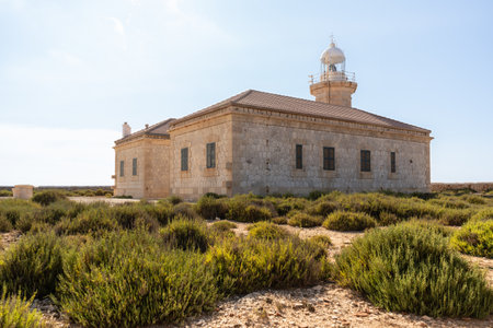 Building exterior of lighthouse of Punta Nati in northern Menorca island, built in 1912, surrounded by bushes in an arid terrain on a warm day. Balearic island tourismの写真素材