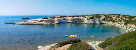 Panoramic view of S'Archittu beach in Sardinia with people enjoying a sunny day. The natural rock arch and clear blue waters create a picturesque coastal scene. Summer tourism concept in Italyの写真素材