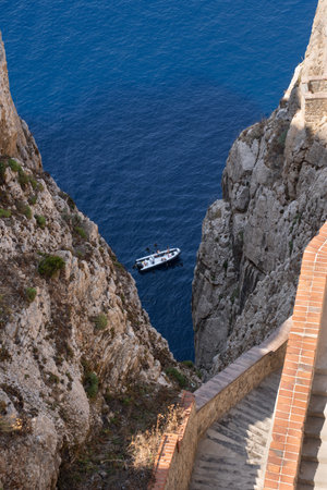 A small boat cruises through the deep blue sea between the rugged cliffs of Neptune's Grotto, captured from the stone staircase leading down in Sardinia, Italyの写真素材
