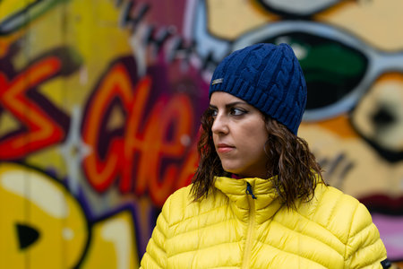 A brunette woman, in her 30s, wearing a yellow puffer jacket and a blue wool hat, looking serious to...