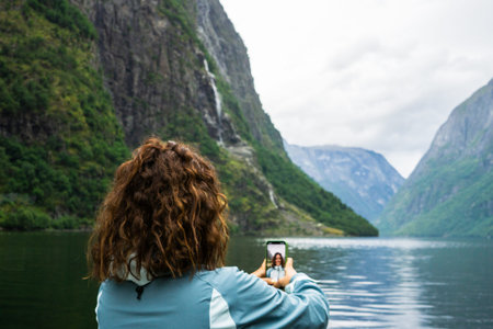 A young woman in her 30s, seen from behind, takes a selfie against the breathtaking backdrop of a Norwegian fjord. The dramatic landscape features steep mountains and calm waters. Active tourism conceptの写真素材