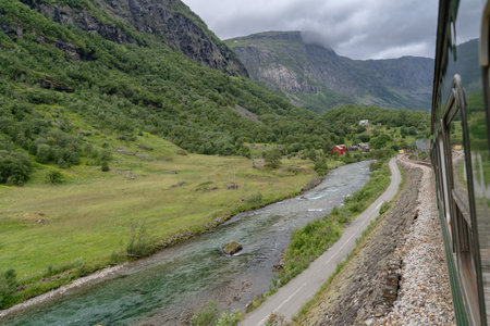 View of the Flam River winding through Flamsdalen Valley, surrounded by lush greenery, mountains, and cloudy skies, captured from the Flamsbana train. Norwegian nature conceptの写真素材
