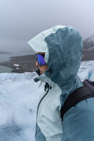 A side portrait of a woman in her 30s wearing a blue hooded jacket, standing on an icy landscape...