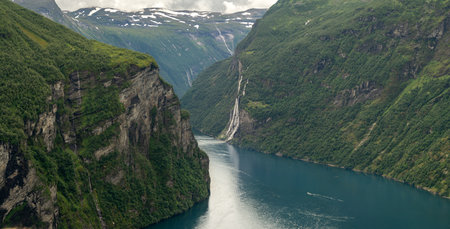 Scenic view of the winding Geirangerfjord in Norway with the famous Seven Sisters waterfalls cascading down steep cliffs, surrounded by lush green nature on a cloudy summer day.の写真素材