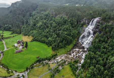 Drone view of Norway's Tvindefossen waterfall falling from a forested mountain with traditional farmhouses by the river, surrounded by lush green fields. Norway tourism conceptの写真素材
