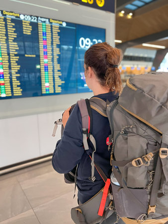 A rear view of a young female traveler carrying a large backpack while looking at the flight...
