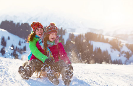 Two joyful young women sledding down a snowy hill in the mountains, laughing and enjoying a sunny...
