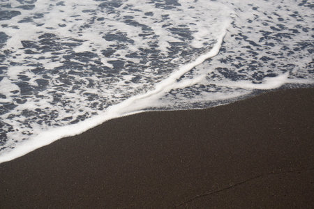 A serene, wide-angle shot of ocean waves with white sea foam breaking onto a pristine dark volcanic sand beach.の写真素材