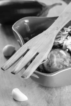 wooden fork, fried eggplant with garlic in a ceramic bowl and raw cloves of garlic and eggplant in black and white, selective focusの写真素材