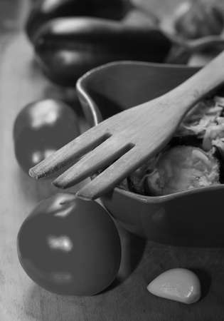 wooden fork, fried eggplant with garlic in a ceramic bowl and raw cloves of garlic, tomatoes and eggplant in black and white, selective focusの写真素材