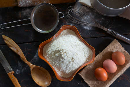 Kitchenware and food at the old home desktop. Wheat flour in a ceramic dish and chicken eggs for the dough preparation. Rustic style. Top view close-up.の写真素材