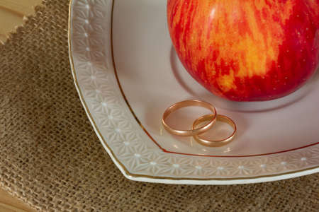 Male and female gold wedding rings and red ripe Apple in a white ceramic saucer on the matting. Closeup top view. Rustic style. Selective focusの写真素材