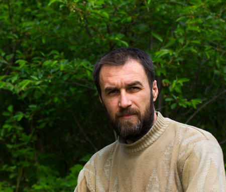 portrait of a pensive adult male with gray hair in his beard against the backdrop of green foliage