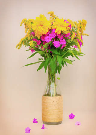 a bouquet of flowers of goldenrod, phlox and lilies in a glass bottle on a pink background. toned photoの写真素材