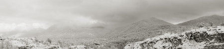black and white panoramic photo landscape of wooded hills in the snow and the clouds and the...