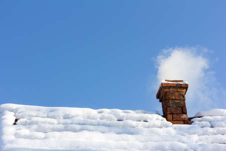 smoke out of a brick chimney on a snowy rooftop on the background of blue skyの写真素材