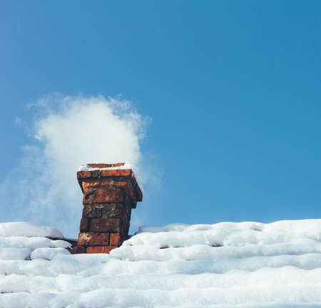 smoke out of a brick chimney on a snowy rooftop home on the background of blue skyの写真素材