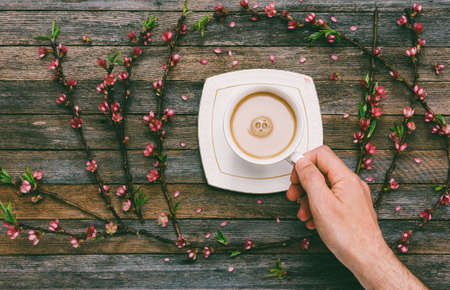 Cup with coffee milk in a male hand on a background of an old wooden table with peach branches with pink flowers, top viewの写真素材