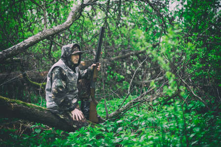 A man hunter with a gun sits on a fallen tree in rainy weather in a spring forest, toned photoの写真素材