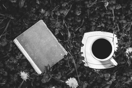 old book and a Cup of hot coffee on a green meadow summer or spring morning top view, black and...