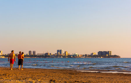 Anapa, Russia - August 11, 2019: Two men with a child walk on a sandy beach on the Black Sea with a view of the city of Anapa on a summer eveningのeditorial素材