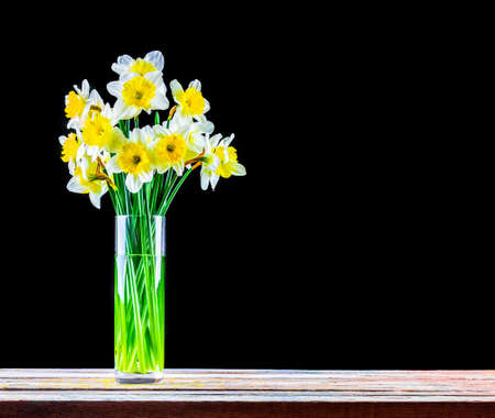 a bouquet of Narcissus flowers in a glass vase on a wooden table on a black background with a copy spaceの写真素材