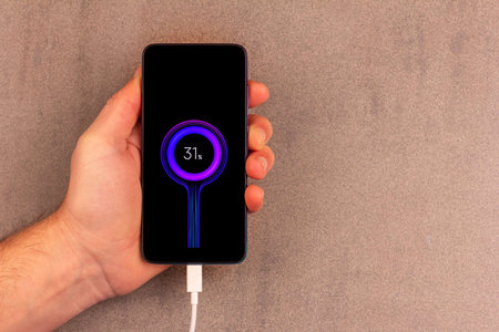 a smartphone with a battery indicator on the screen with a white charging wire in a male palm on a...