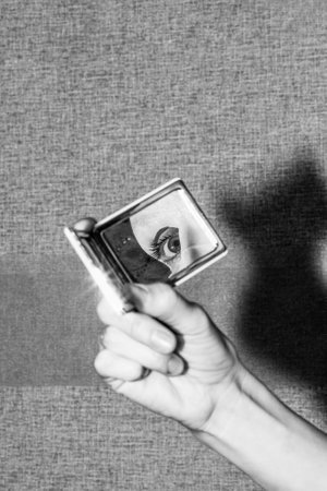 mirror in hand and reflection of the eye and moisturizing mask on the face of a young woman, black and white photoの写真素材