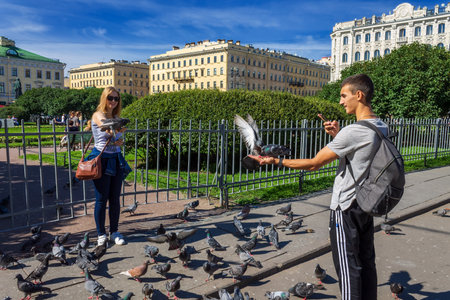 Saint Petersburg, Russia - August 9, 2019: a beautiful guy and a girl feed pigeons with their hands and shoot a video on a smartphone on a summer dayのeditorial素材