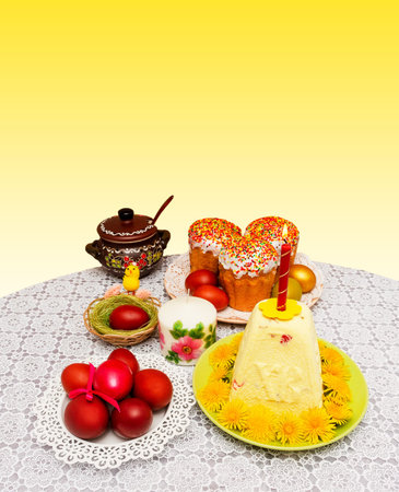 Cakes and painted eggs with a yellow chicken in a nest on a table on yellow and white