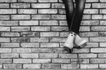 crossed legs of a young girl in jeans and orange galoshes sitting on a brick wall against a brick wall background with copy space, black and white photoの写真素材