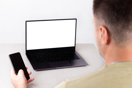 a man with a phone in his hand is sitting in front of a laptop with a white mockup on the monitor,...