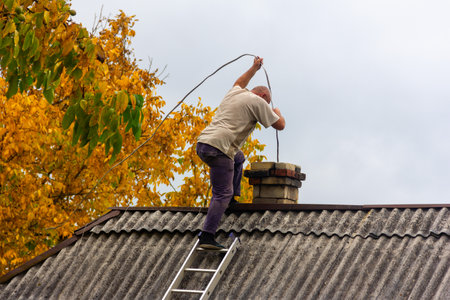 a male chimney sweep cleans the chimney from soot on the roof of a village house, preparation for the heating seasonの写真素材