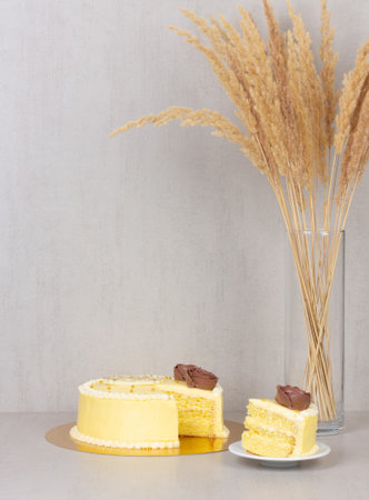 saucer is a cut piece of cake from a yellow cream cake with chocolate roses and a bouquet of dried reed flowers in a glass vase on a gray background with copy spaceの写真素材