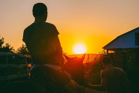 a young hostler leads a horse by the bridle with a man and a child sitting on it against the background of a summer sunsetの写真素材