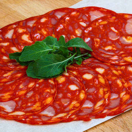 A close-up overhead view of thinly sliced, vibrant red chorizo sausage arranged in a circular pattern, topped with a small cluster of fresh green arugula.の写真素材